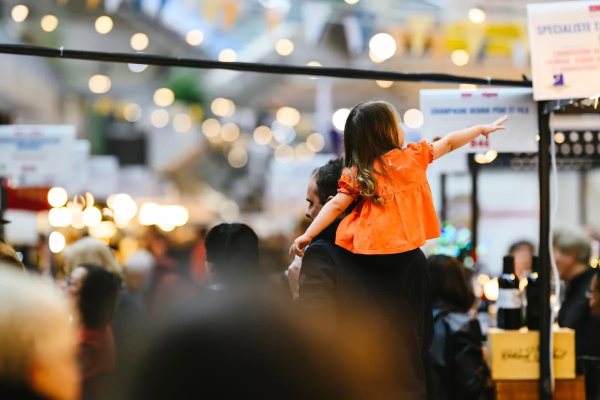 Père portant sa fille sur ses épaules dans l'ambiance festive et conviviale d'un salon Mer et Vigne, entre guirlandes lumineuses et stands de vins et champagnes
